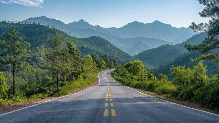 Winding mountain road through a green forest with distant blue ridges under a clear sky.