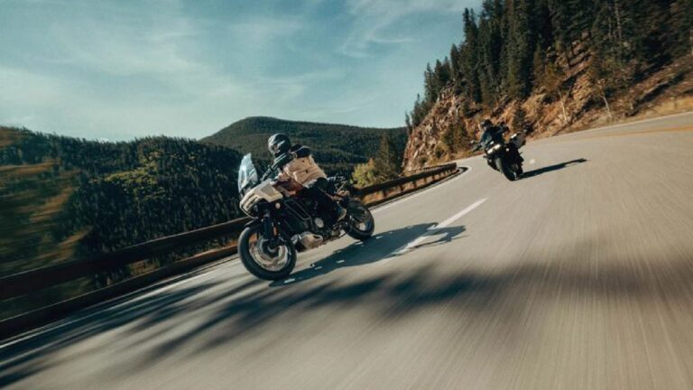 Two motorcyclists lean into a sharp bend on a winding mountain road, forested slopes on one side and guardrail on the other.