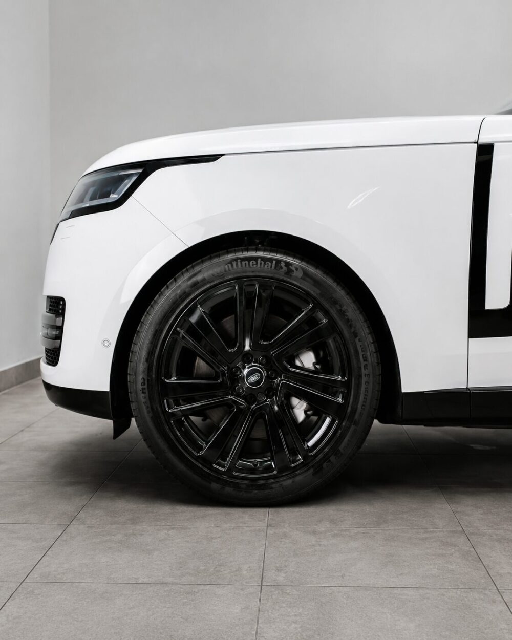 Close-up of a white car's front left wheel and fender in a showroom setting.Large glossy black alloy wheel with Continental tire visible on a white vehicle.