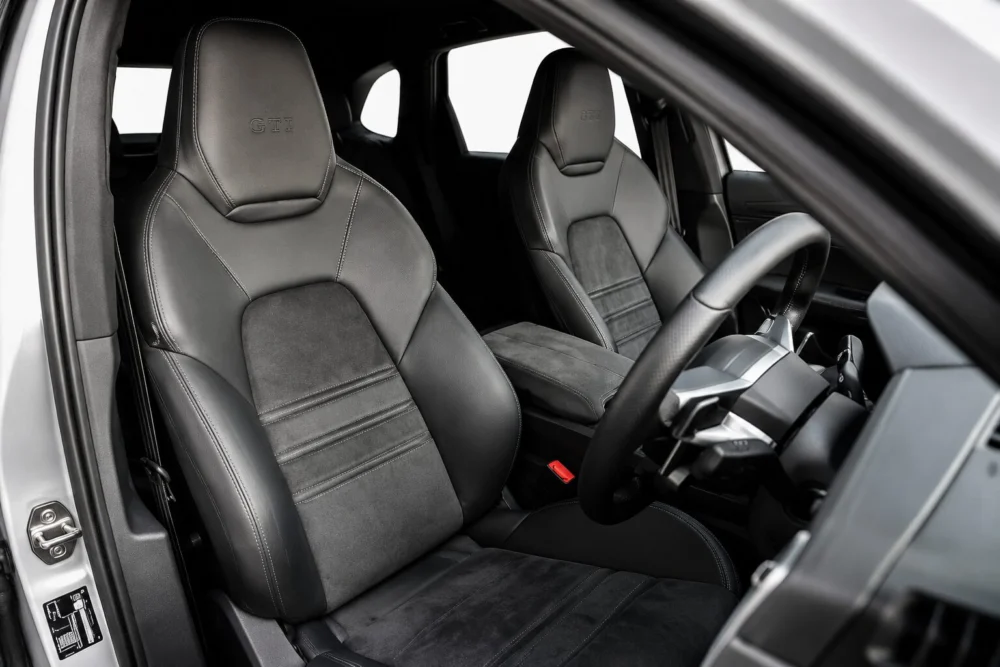 Interior of a right-hand-drive car showing two sporty black leather and suede bucket seats with GTI embroidery and a steering wheel nearby