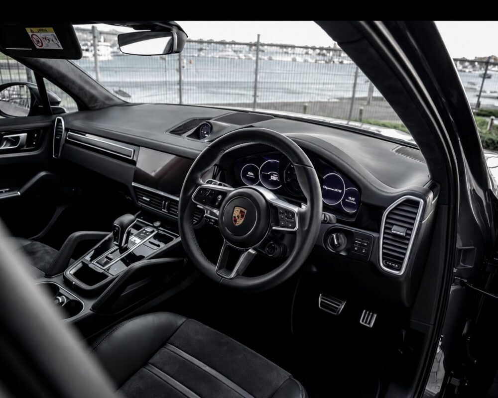 Luxurious Porsche interior with a leather-wrapped steering wheel, digital gauges, and a sleek center console near the driver’s seat, by a waterfront view outside the window.