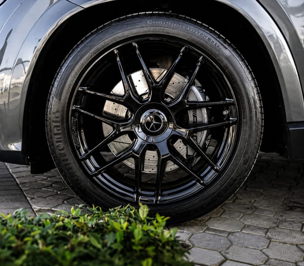 Close-up of glossy black multi-spoke car wheel with silver brake disc and Mercedes center cap on a dark tire surface.