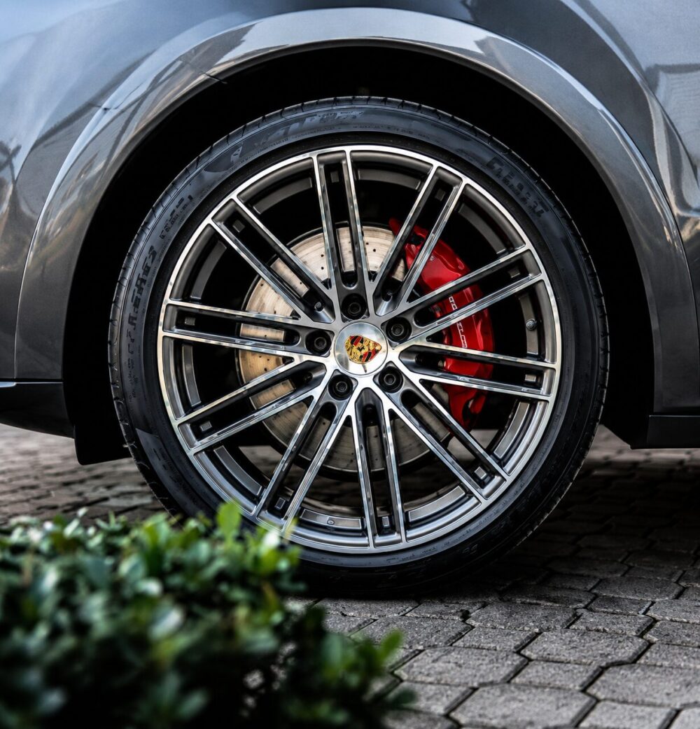 Close-up of a Porsche alloy wheel with multi-spoke design, red brake caliper, and drilled rotor behind a tire on a paved surface.
