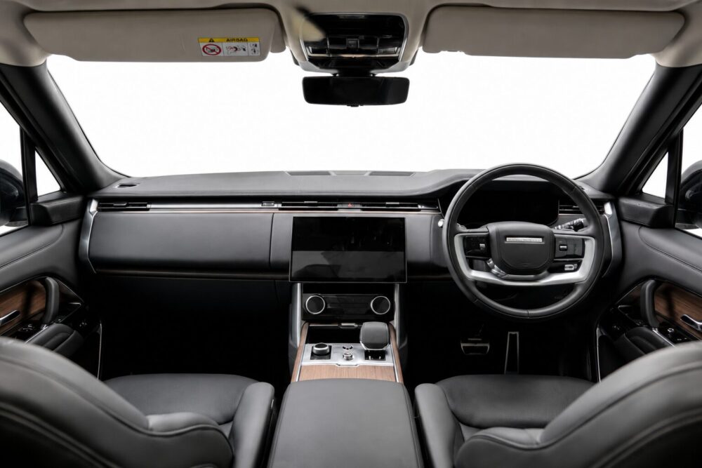 Interior of a car cabin showing a large central touchscreen, wood trim, and a right-hand steering wheel with controls on a black dashboard.