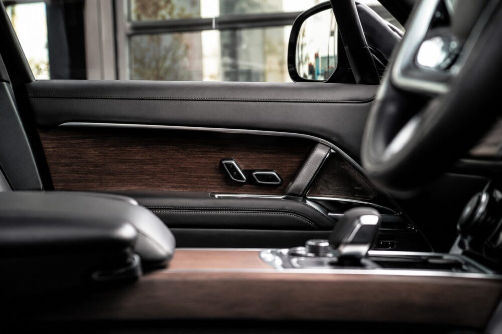 Close-up of a luxury car door interior: dark leather, wood veneer panel, and chrome control switches near the armrest.