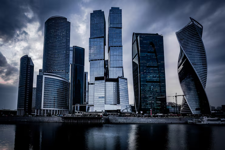 Skyline of a modern glass skyscraper cluster along a riverbank under a cloudy sky, with a distinctive twisting tower on the right side.